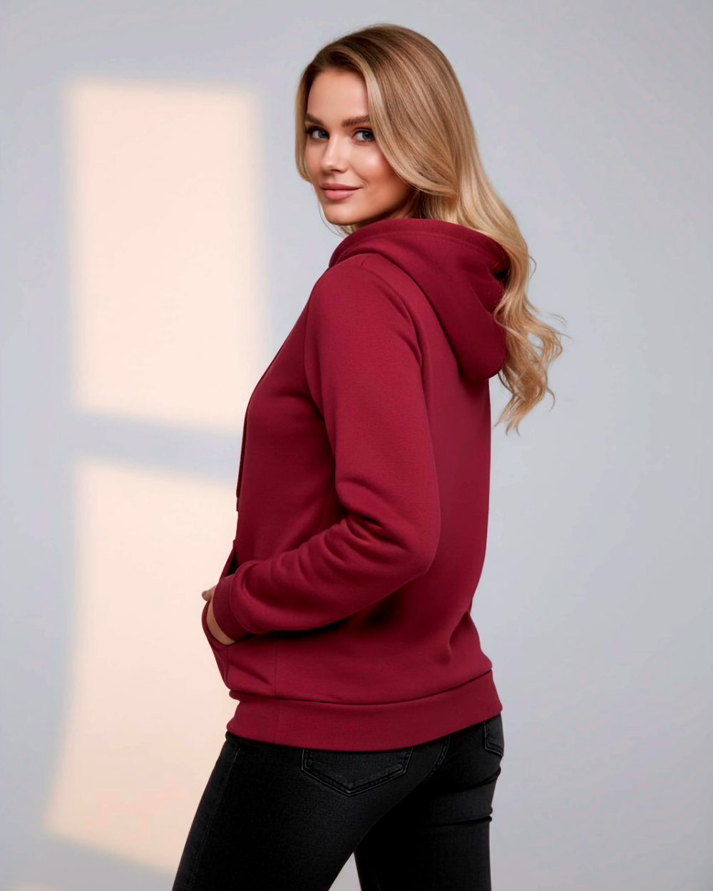 Modelka ujęta z półprofilu, tyłem do aparatu, prezentuje tył bordowej bluzy z założonym kapturem. Jedna dłoń schowana jest w bocznej części przedniej kieszeni.