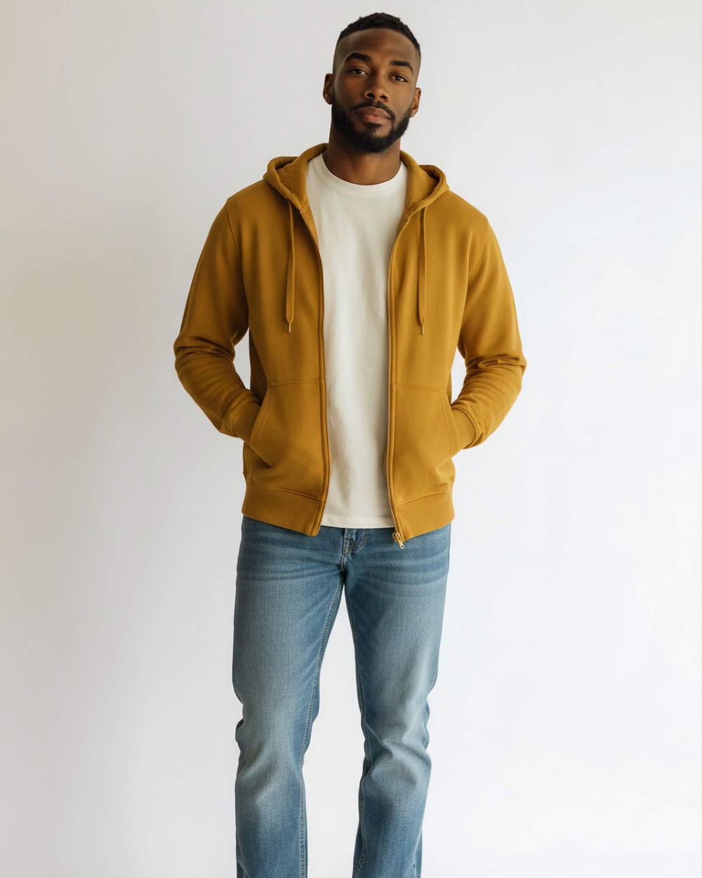 Mężczyzna stoi w jasnym pomieszczeniu, na jasnym tle, ma na sobie klasyczną bluzę na suwaku, z kapturem i kieszeniami w których trzyma dłonie. Bluza ma modny musztardowy kolor, jest rozsunięta, widać biały T-shirt. Stylizację dopełniają klasyczne, jasne jeansy.